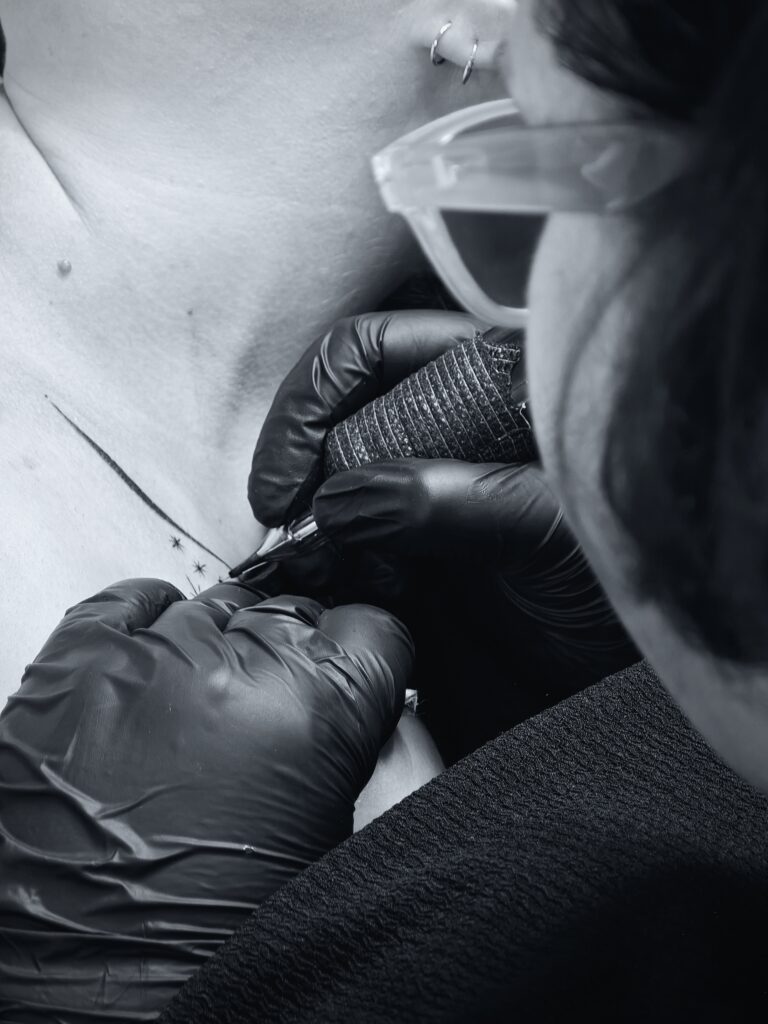 Schwarz-graues Bild, Ansicht über die Schulter der Künstlerin bei der Ausarbeitung eines Fineline Tattoos am Schlüsselbein